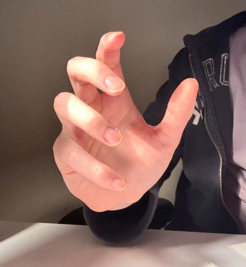 a closeup image of a hand with long fingers extended towards the camera. the lighting is soft and natural, coming from the left side of the frame. the background is a blurred white surface and part of a black jacket.