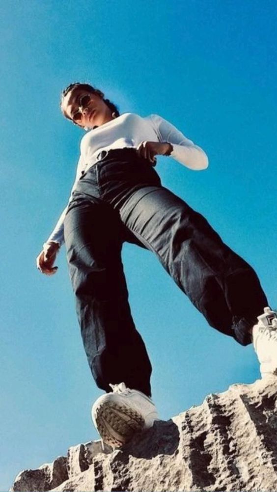 a young woman in a white shirt, blue jeans, and white sneakers stands with one foot on a rock, posing for a picture taken from a low perspective against a clear blue sky.