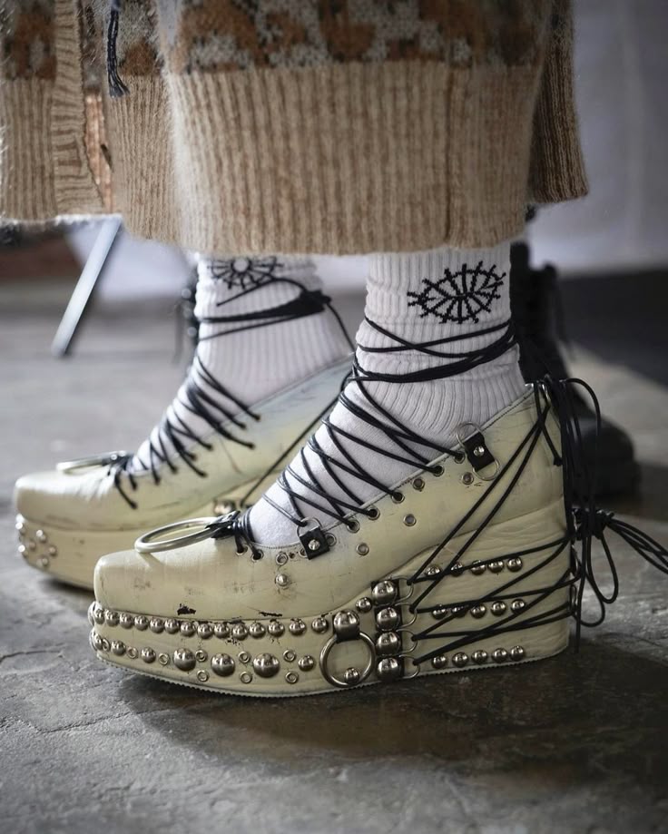 a closeup shot showcases a pair of platform shoes adorned with metal studs and decorative rings, worn with patterned socks. the white socks feature a distinctive eye pattern, and the shoes are secured with black laces that wrap around the ankles. the shoes have a wedged heel, adding to their distinctive style.