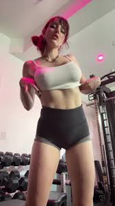 a young woman with vibrant red hair is captured in a gym setting, showcasing her physique.  she's wearing a white cropped top and dark athletic shorts. she's positioned in front of weight machines, dumbbells are visible in the background, suggesting a fitness or workout context. the lighting enhances the woman's features and the overall mood of the image. the overall impression is one of fitness and attractiveness.