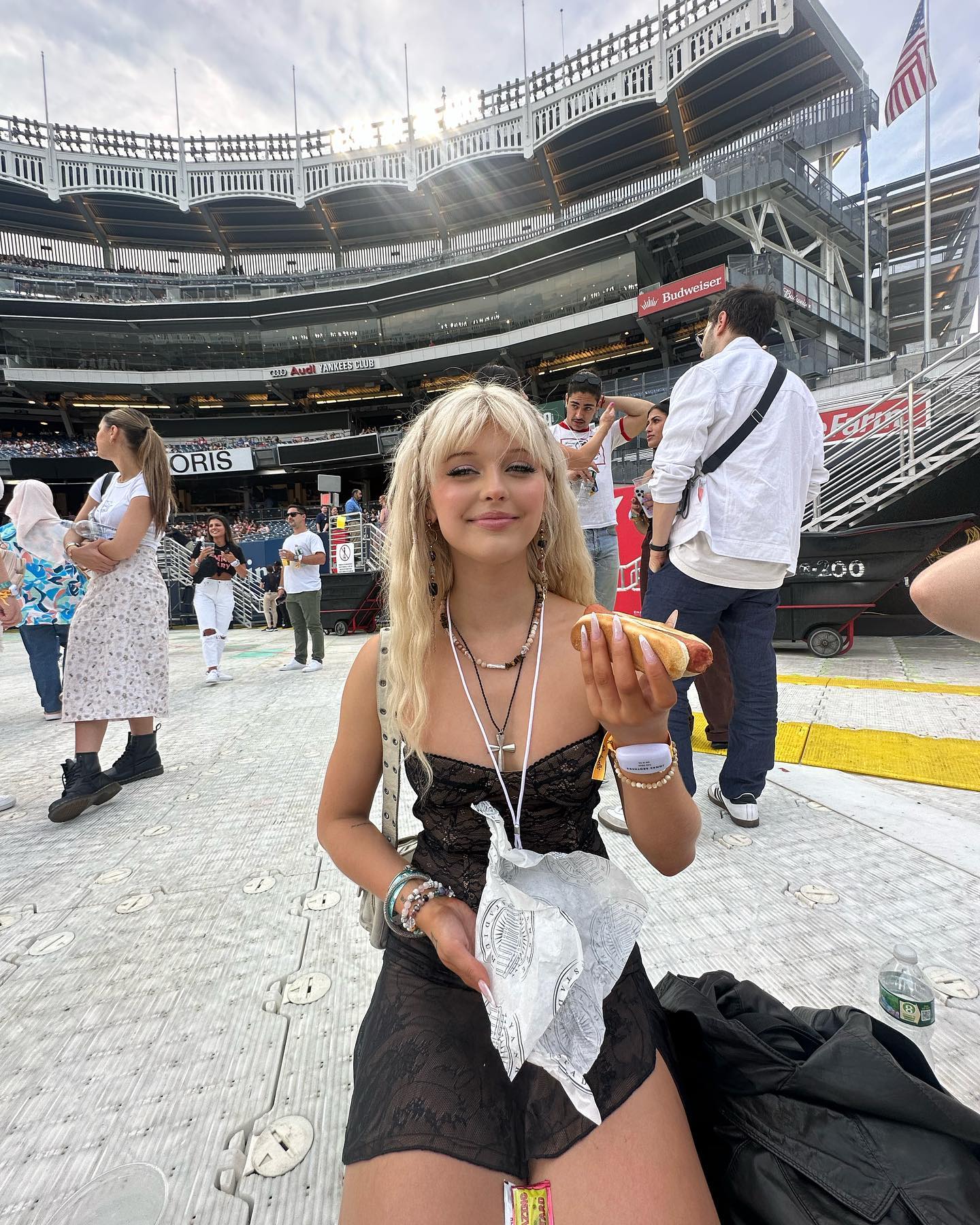 a young woman with blonde hair is sitting in a stadium holding a hot dog and a paper napkin. she is wearing a black dress, necklaces, and bracelets. behind her, there is a crowd of people, a budweiser sign, and the stadium structure. a water bottle is visible on the ground near her. the stadium appears to be a baseball stadium, possibly yankee stadium. she is enjoying a hot dog at a baseball game.