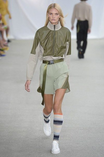 a female model walks down a runway wearing a stylish outfit. the outfit consists of a light olive green and beige color blocked jacket, light green shorts, a matching olive green belt, and striped kneehigh socks with white sneakers.  the model has blonde hair and is walking confidently. another person is visible in the background, out of focus. the overall setting appears to be a fashion show.