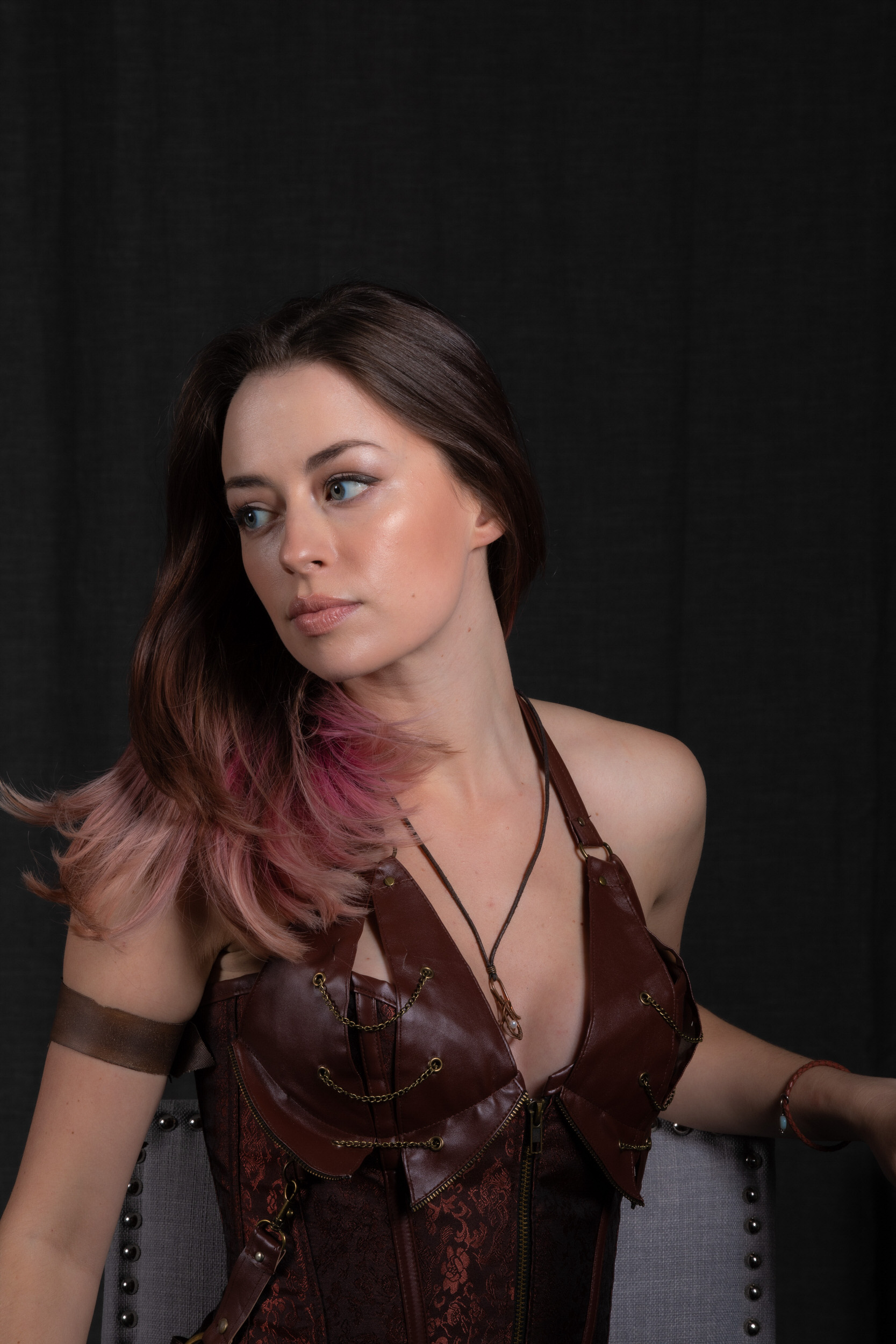 a woman with brown and pink hair and a brown steampunkstyle top sits facing left with her head turned slightly away from the camera. she wears a leather and metal arm band on her left arm.  the background is black, and the woman sits on a gray chair.