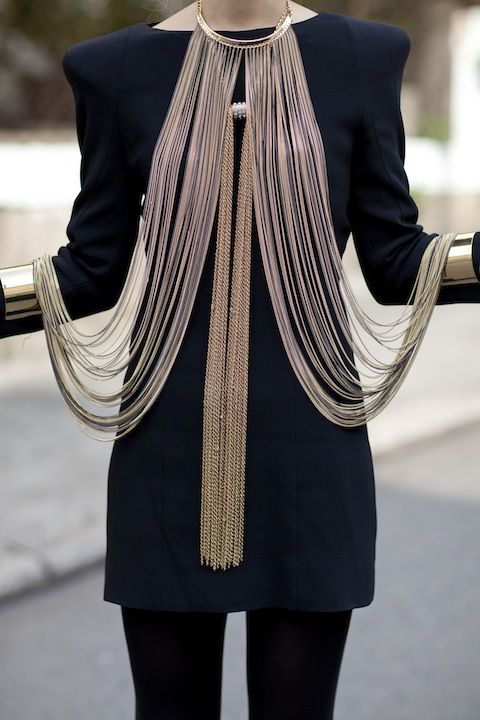 a woman is wearing a black dress with a striking gold necklace and arm cuffs. the necklace is comprised of layered chains of varying lengths, creating a dramatic cascade effect. the gold arm cuffs are wide and bold, complementing the necklace. the woman also appears to be wearing black tights. the image has a highfashion and elegant vibe.
