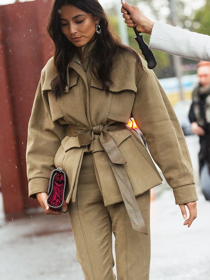 a stylish woman walks down a city street in the rain. she is wearing a beige suede pantsuit with a belted jacket and matching trousers.  the jacket has flap pockets and a relaxed fit. she carries a dark burgundy velvet handbag and wears simple silver hoop earrings. the overall look is autumnal and chic.  part of another person's arm holding an umbrella is visible in the upper right corner.