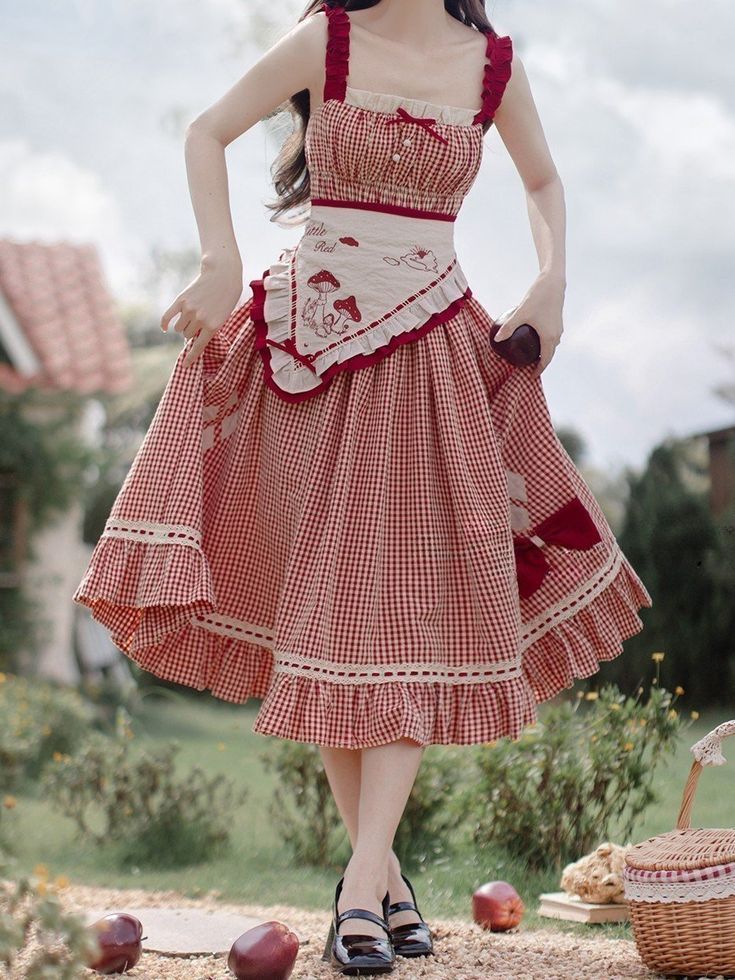 a woman in a red and white gingham dress is standing in a field, holding up the skirt of her dress. the dress has a sweetheart neckline, a ruffled hem, and a white apron with embroidered mushrooms on it. she is wearing black mary jane shoes and is holding a red apple in her right hand. there is a picnic basket on the ground behind her and a few apples scattered around her feet. the background is a field with green grass and a blue sky with some clouds. the lighting is natural daylight and the overall feel of the image is sunny and romantic.
