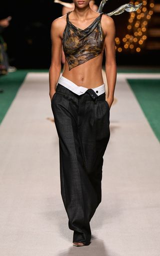 a female model in a crop top and pants walks the runway during a fashion show. she is wearing a black crop top and black pants with a white belt. she is also wearing her hair up in a ponytail.