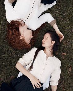 the image shows two women laying on a grassy field, their heads close together and their bodies forming a heart shape. one woman has long red hair and the other has long brown hair braided down her back. both women are wearing white shirts. the woman with the brown hair is also wearing a black skirt. the image has a soft, romantic feel, with natural light creating a warm and inviting atmosphere.
