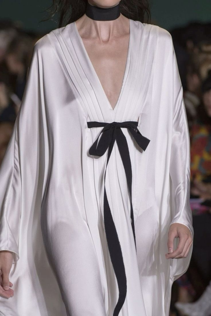 a woman in a white silk dress with a black bow tied at the waist and a black choker. she is walking down a runway, her dress billowing around her.