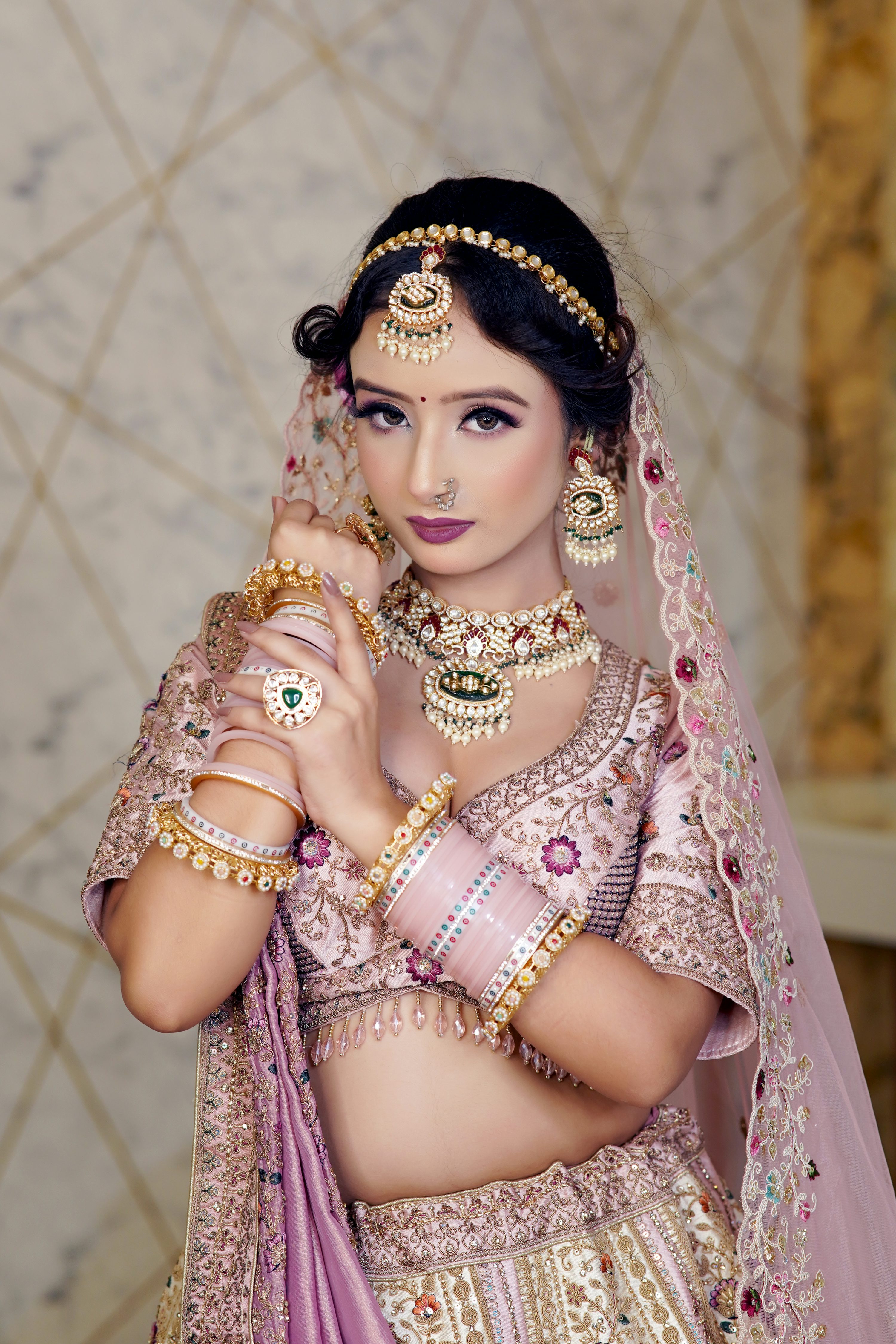 the image showcases a stunning indian bride adorned in a beautiful light pink and gold lehenga choli. she's wearing elaborate traditional jewelry, including a maang tikka (head jewelry), a necklace, earrings, bangles, and a nose ring. her makeup is impeccable, enhancing her features. the bride's pose and expression project elegance and grace.  the backdrop appears to be a textured wall providing a soft, neutral contrast to her vibrant attire. the overall feel is one of luxury and traditional indian bridal beauty.