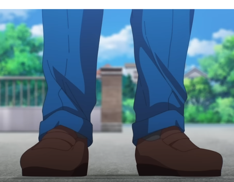 this is a low angle shot of a man wearing blue trousers and brown shoes standing on a pavement. there are trees, a brick wall, and buildings in the background.