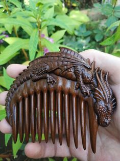 a hand is holding a brown wooden comb, intricately carved with a dragon figure on the top. the comb is in focus, with a background of green plants.