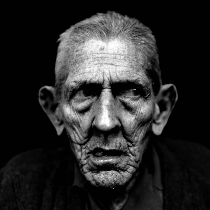 a black and white closeup portrait of an elderly man with a heavily wrinkled face. he has a serious expression and is looking slightly to the side. the lighting is dramatic, highlighting the textures and contours of his face. the background is dark and black, emphasizing the man's features.