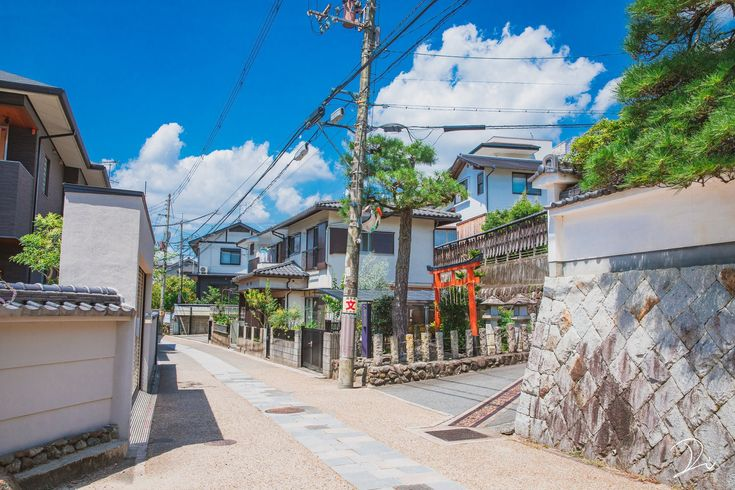 a street view in a residential area with buildings, houses, and a torii gate under a blue sky with clouds. a utility pole stands in the street, and a wall is visible on the side.