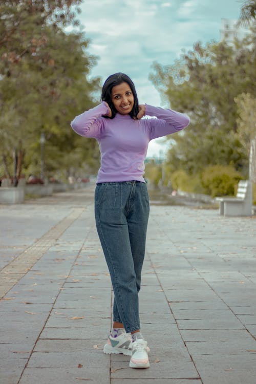 a woman is standing on a paved walkway outdoors. she's wearing a longsleeved purple top, dark wash jeans, and white sneakers. she has her hands up near her neck and is smiling. the background includes trees, a bench, and a cloudy sky.