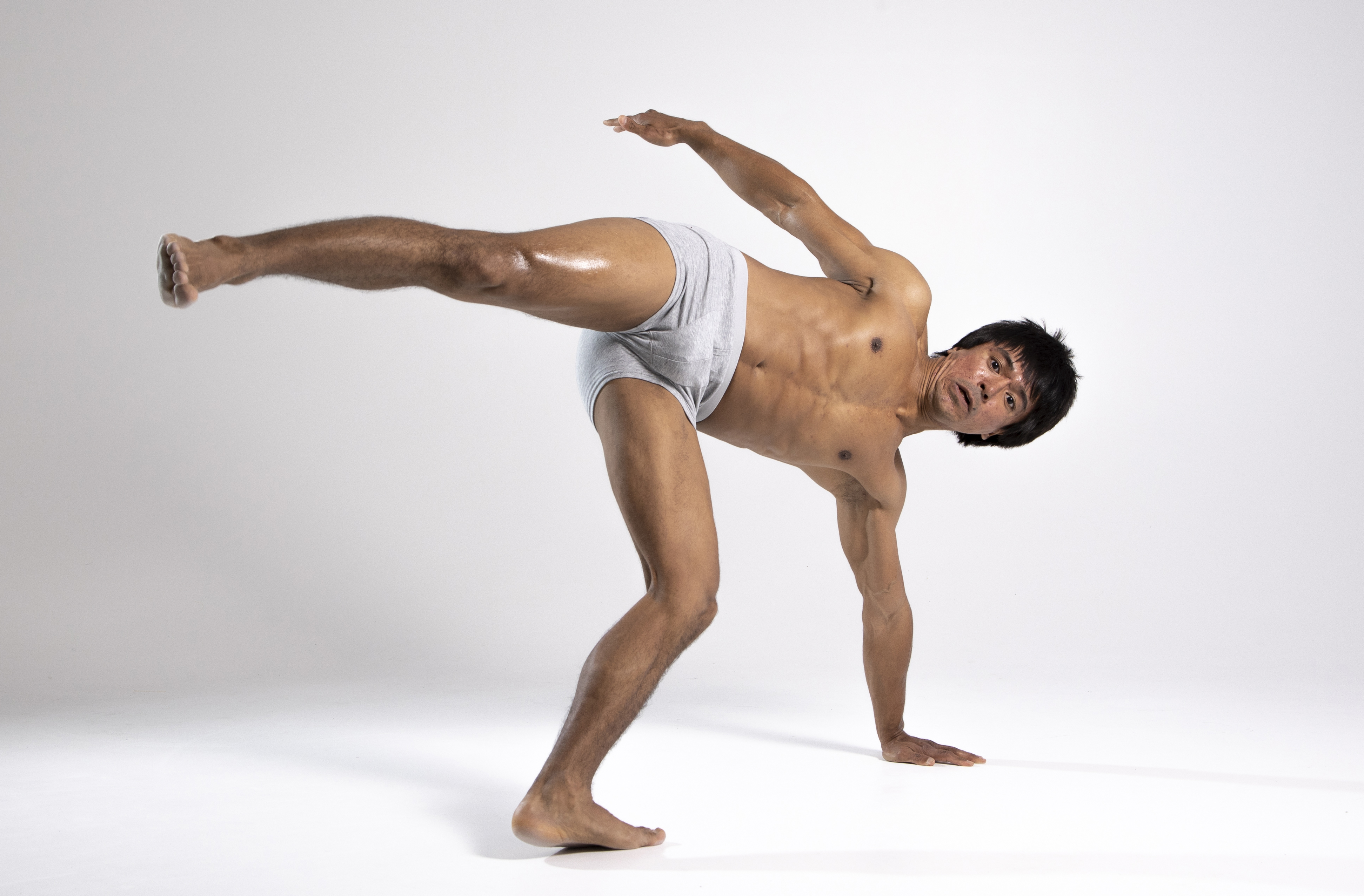 a shirtless man in underwear is posed on a white background, balancing on one hand while extending one leg and the other arm for balance. he appears to be executing a challenging physical exercise or a martial arts move.