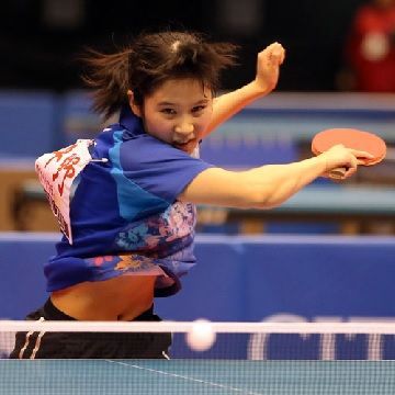 a young woman, wearing a blue and white table tennis uniform, is midserve, holding her paddle in her right hand and concentrating intensely on the ball, her eyes fixed on the trajectory of the ball. she has a headband in her hair and a faint smile on her face, suggesting her joy and excitement for the sport. the background is blurred, highlighting the athlete's action and emphasizing the focus on the player. the image captures the intensity and concentration of a table tennis competition.