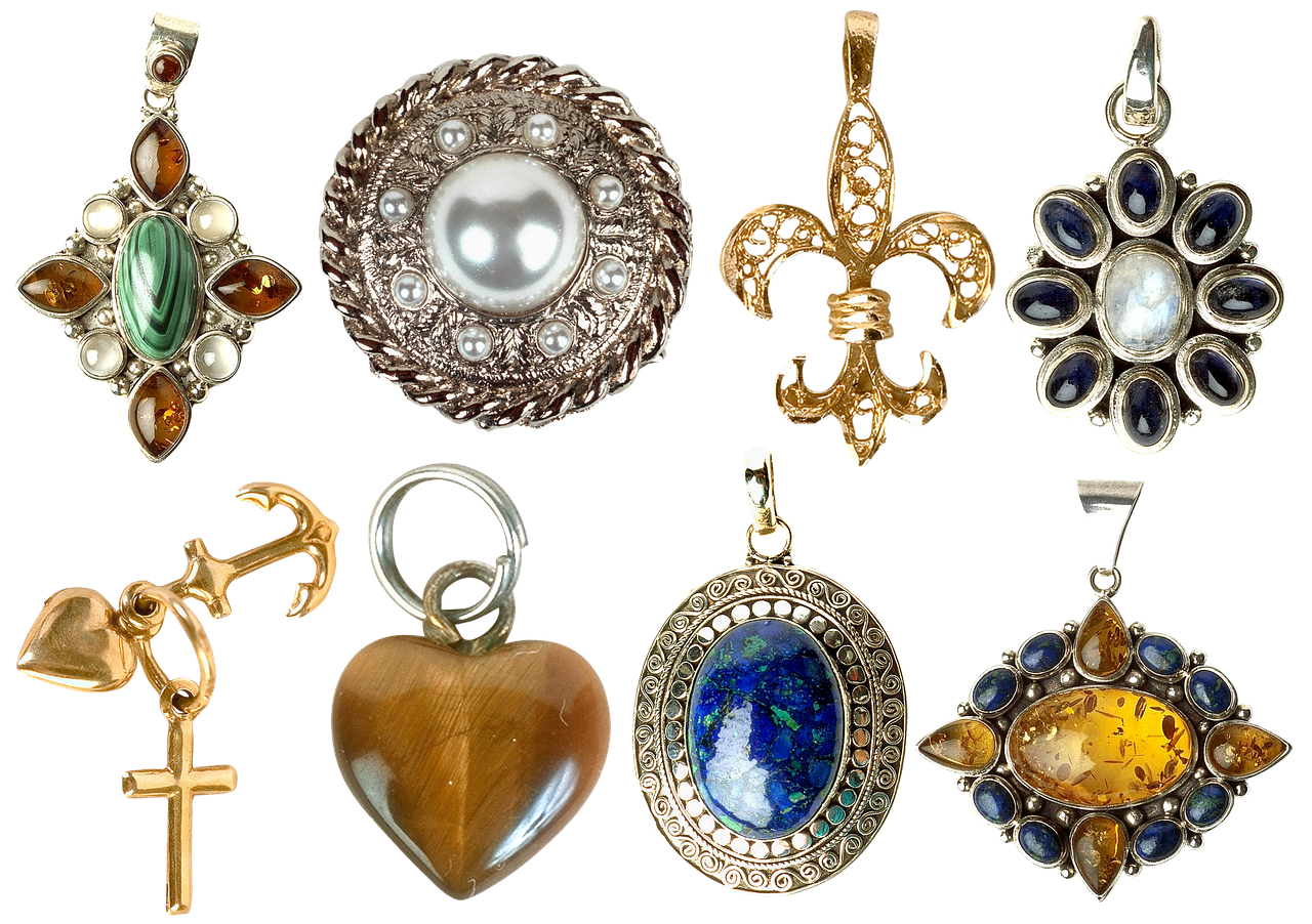 the image displays a collection of jewelry pendants against a black background. the pendants feature various designs, including floral, geometric, and symbolic motifs, such as a fleurdelis, an anchor with a cross, and a heart. a variety of gemstones are used, including malachite, amber, lapis lazuli, and moonstone, along with pearls and tiger's eye. the metals used are gold and silver.