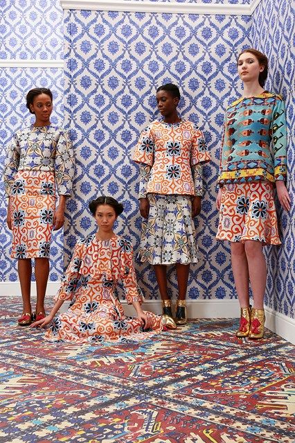 four female models pose in a studio setting against a backdrop of blue and white patterned wallpaper. they are wearing matching outfits in a variety of bright colors with a similar style reflecting a mix of ethnic inspired patterns with both floral and geometric designs. the outfits include skirts and tops featuring  vibrant colors and intricate prints. the models are positioned  in a row.  the floor is covered with a similarly patterned carpet creating a unified and visually striking effect. the overall aesthetic suggests a fashion photoshoot or runway presentation focused on a collection of unique printed clothing.