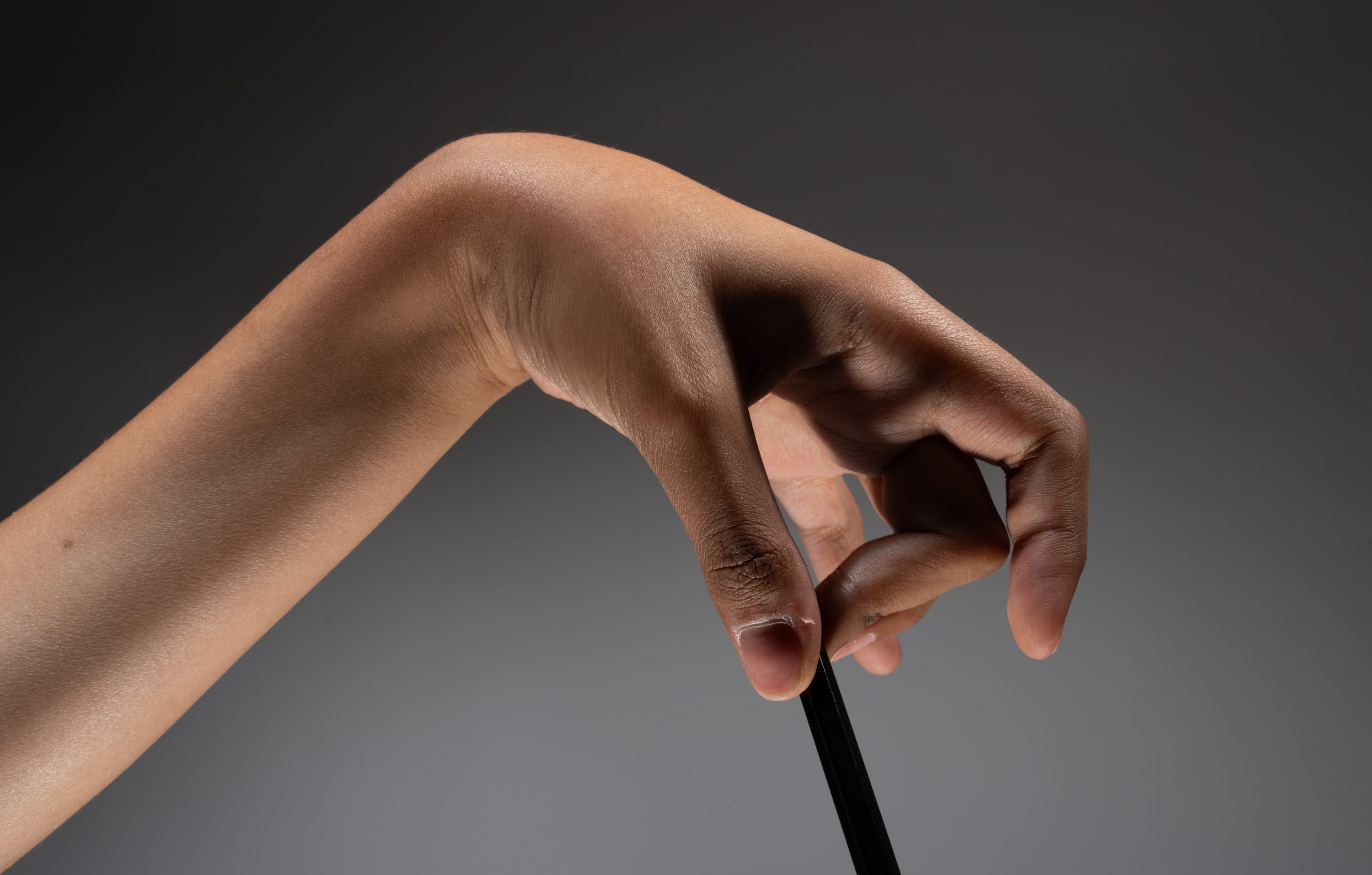 a closeup image of a hand delicately gripping a black pencil against a neutral gray backdrop. the hand is positioned in a clawlike grip with the pencil held vertically. the studio lighting highlights the texture and subtle color variations of the skin.