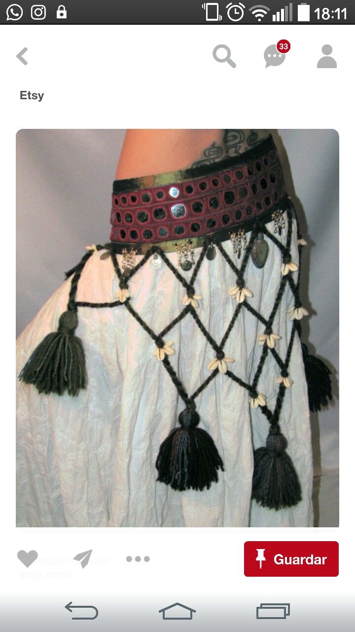 the image shows a closeup view of a belly dance costume's lower half.  the costume features a burgundy waist belt with circular cutouts, adorned with metallic accents. from the belt, a netlike structure hangs down, decorated with small white shells and dark green tassels. the skirt itself is a flowing offwhite fabric.  the overall style is ethnic and bohemian, suggesting handcrafted or artisanmade clothing. the picture is likely taken from an etsy listing, as indicated by the "etsy" label at the top.  the background is blurry and out of focus, keeping the attention on the costume detail.