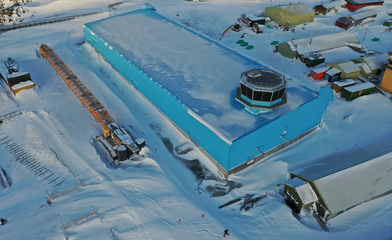 an aerial photo of a snowcovered airport with a large blue building with a control tower on top. the airport is located on a snowcovered landscape. there is a large yellow machine in the bottom left corner of the image.