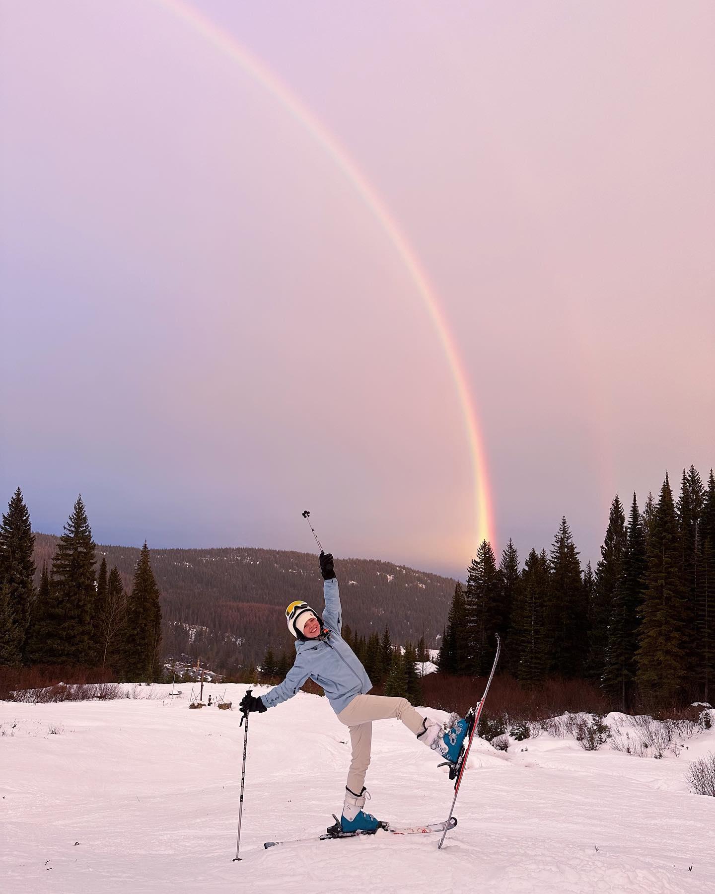 the image shows a woman on skis, striking a celebratory pose with her arms raised in the air.  she's positioned on a snowy mountain slope with a backdrop of a beautiful, partially visible rainbow arching across a pastelcolored sky at sunset or sunrise. a line of dark evergreen trees forms a picturesque horizon. the scene conveys a feeling of joy and freedom in a stunning winter setting.