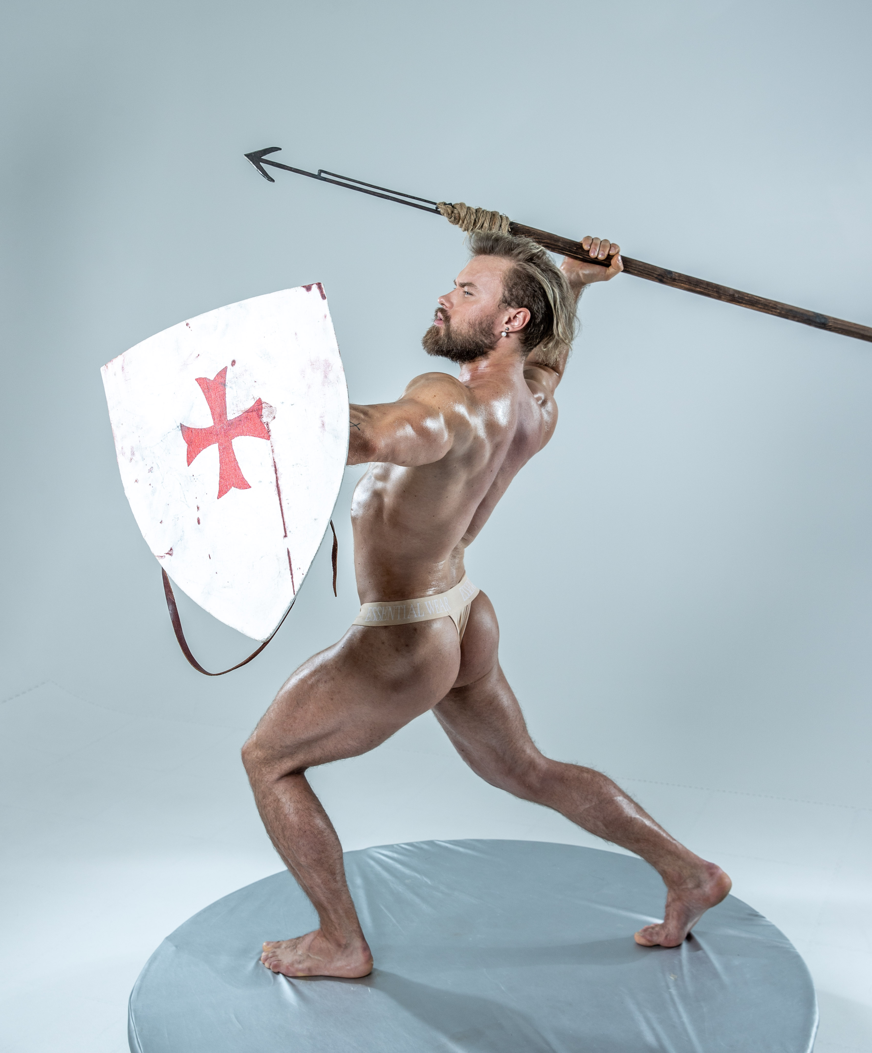 a muscular man, posed as a warrior or knight, stands in a dynamic stance against a light gray studio background. he holds a wooden spear aloft in his right hand and a white shield, emblazoned with a red cross, in his left. his body, oiled and glistening, suggests strength and power. the image evokes themes of battle, history, and medieval combat.  the artificial studio lighting highlights his physique and the textures of his weapons.