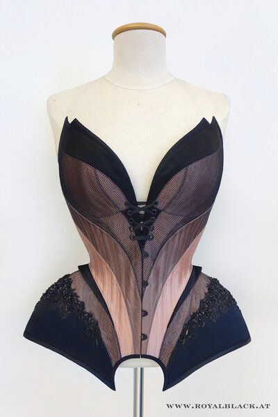 an image showcases an ornate corset displayed on a mannequin against a plain white background. the corset features a complex design with alternating panels of brown and pink hues layered with black mesh. black fabric accents the top and bottom edges, with intricate black lace detailing at the bottom. the corset is cinched at the front with a laceup closure and button details. the website address "www.royalblack.at" is visible at the bottom of the image.