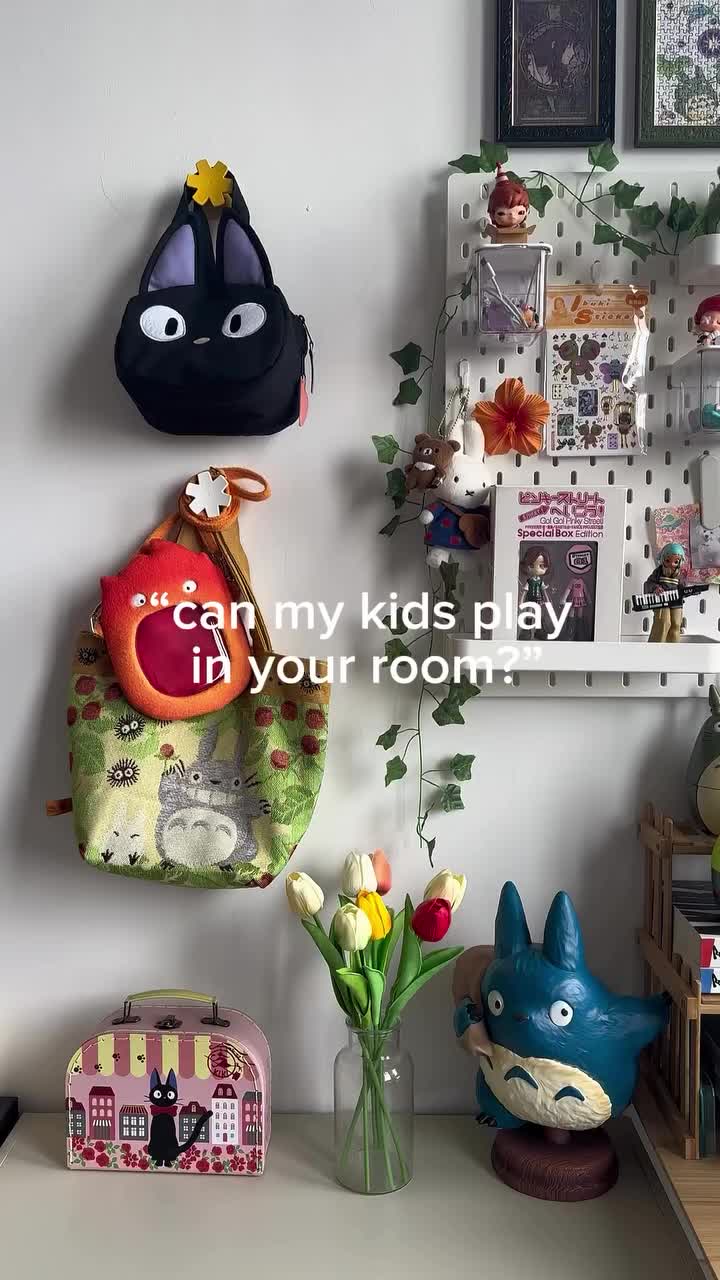 a shot of a decorated wall and shelf in a room. the wall has two hanging studio ghibli themed bags, one with totoro and the other jiji the cat from kiki's delivery service. the shelf to the right is decorated with toys and figurines, and a fake ivy is growing around the edges. on the desk underneath is a jiji carrying case, a glass vase with tulips, and a totoro figurine. text overlayed on the shot asks "can my kids play in your room?"