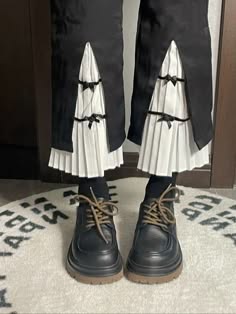 a person is wearing a black and white lolita outfit. the skirt is black with a white ruffle trim. the shoes are black laceup boots. the person is standing on a white rug with the word "japan" on it. the person's legs are crossed at the ankles. the person's feet are turned inward. the photo is taken from a high angle. the photo is well lit. the background is blurry.