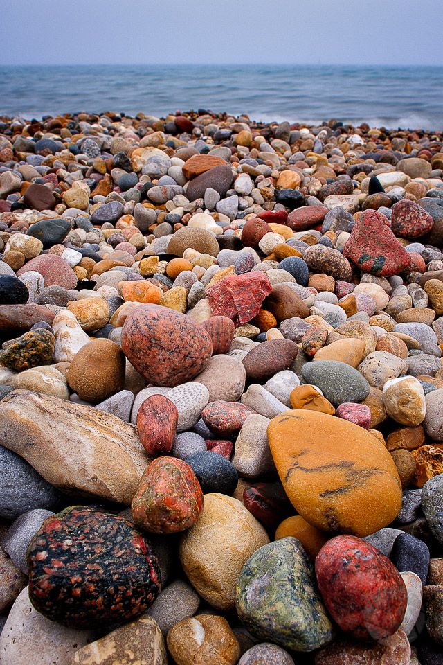 a closeup view of a variety of smooth, colorful rocks and pebbles, laying on a beach. the ocean water is in the background. the rocks are arranged in a random pattern, with many different colors and textures. the ocean is calm and blue. the image is a beautiful representation of nature's beauty.