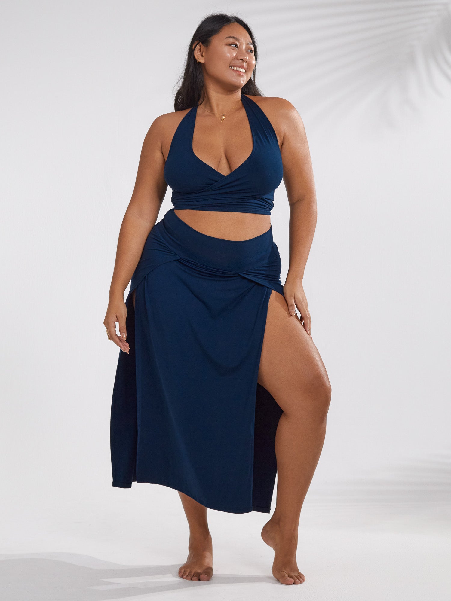 a plussize model is showcasing a stylish navy blue twopiece swimsuit. the set includes a halter neck top and a highwaisted maxi skirt with a thighhigh side slit. the model is standing against a white backdrop, with soft lighting illuminating her.  the overall style suggests beachwear or summer fashion. the image focuses on body positivity and inclusivity in fashion.