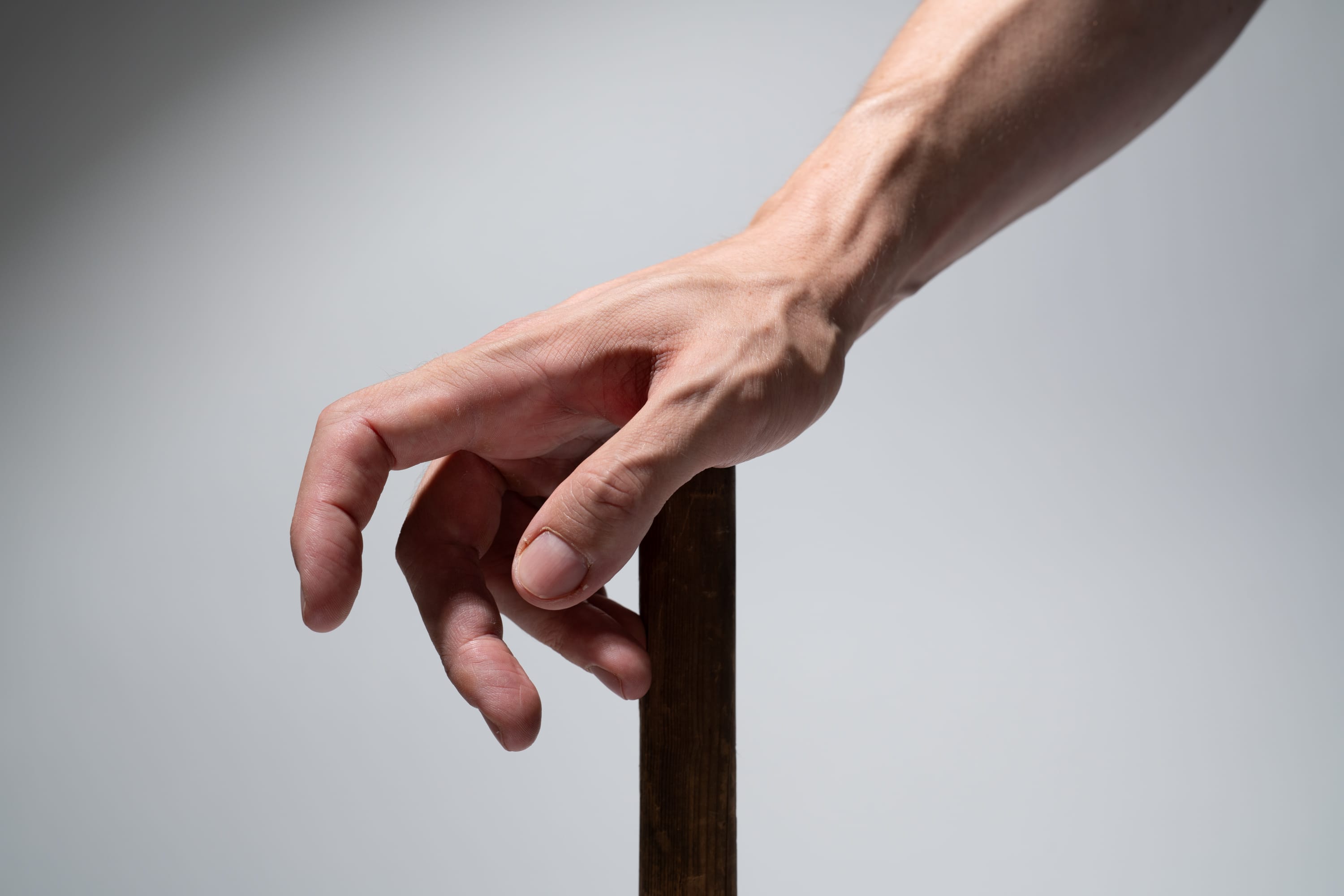 a man's hand is shown gripping a dark brown walking stick against a gray background. the hand is illuminated with studio lighting, highlighting the veins and details of the skin.