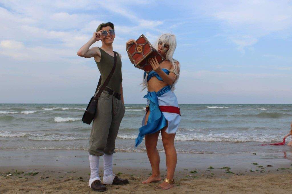 two cosplayers dressed as milo thatch and kida from the disney movie atlantis are standing on a beach in front of the ocean. milo, on the left, is wearing glasses, a green tank top, khaki pants, white socks, brown shoes, and a shoulder bag. he is holding up one hand as if looking into the distance. kida, on the right, is wearing a blue and red costume with white hair and gold accessories. she is holding and looking at a brown book with a red swirl on it. the sky is partly cloudy.