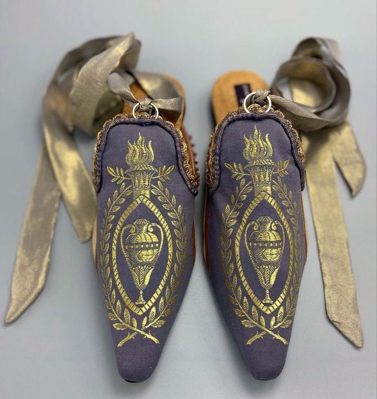 a pair of slipon mule shoes are displayed on a gray surface. the shoes are predominantly a muted purple color, adorned with golden designs. these designs include patterns resembling torches, laurels, and vases. each shoe has a golden ribbon attached via a ring. the ribbons are loosely tied and arranged to the sides of each shoe. the mules appear to be made of fabric, with decorative trim bordering the top of the shoes.