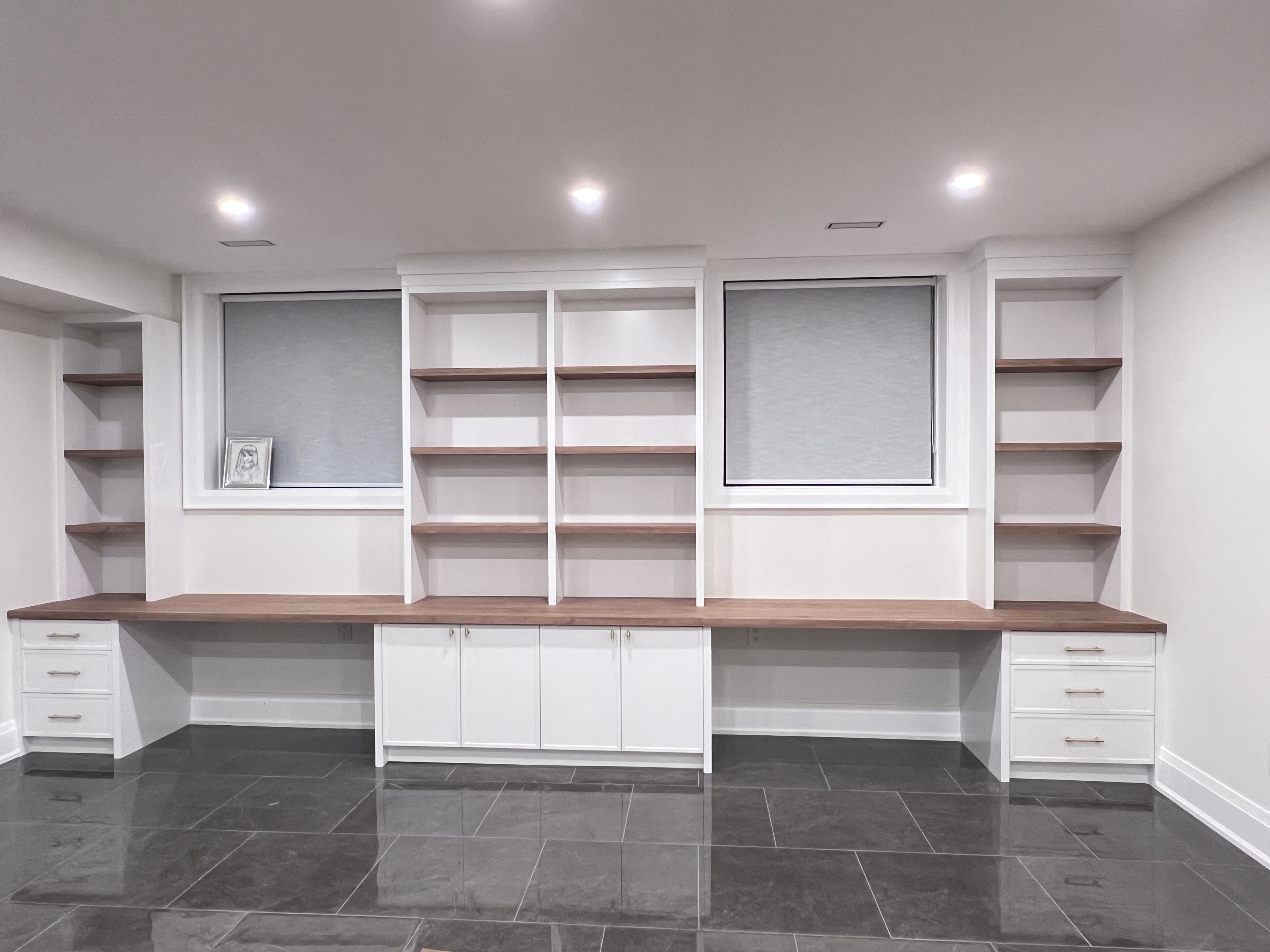 he image shows a newly renovated basement home office featuring a long, builtin desk with a walnut countertop.  the desk is flanked by white cabinets with drawers and additional shelving units on either side.  the shelving units offer ample storage space. two windows with lightgrey blinds are set within the wall unit. the room has recessed lighting and dark grey tiled flooring, contributing to a modern and contemporary aesthetic. the overall design is clean and sophisticated.