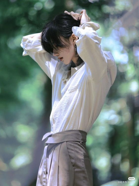 a young man with long black hair and tattoos is shown tying his hair back, wearing a white shirt and grey wideleg pants. the background is a softfocus green, suggesting a natural setting.