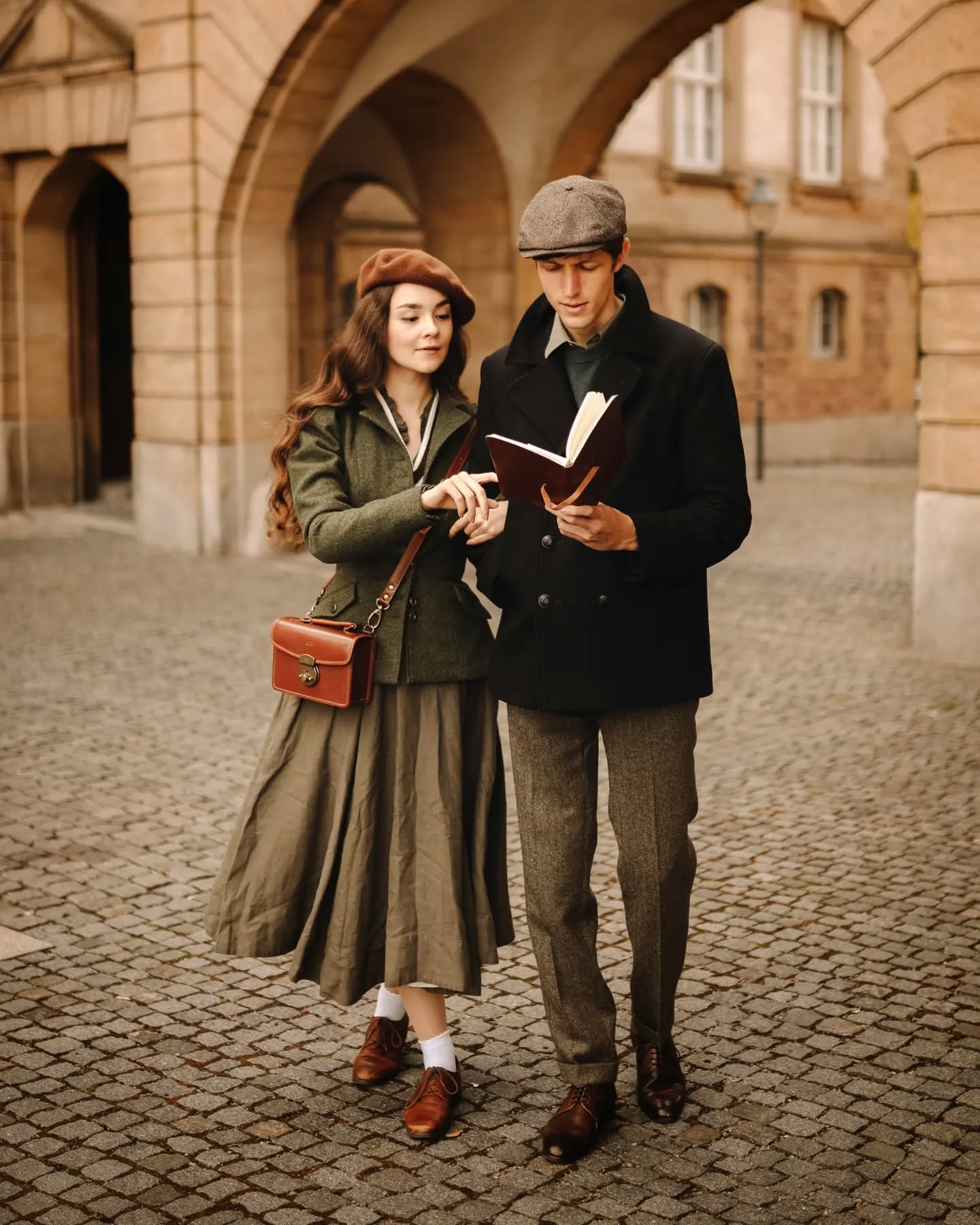 a vintagestyle photograph of a young couple dressed in 1920s or 1930s attire.  the woman wears a green dress, brown hat and brown shoes, and carries a small brown leather handbag. the man wears a dark coat, cap, and brown trousers.  they are standing on a cobblestone street under archways of a stone building, and are sharing a book together. the overall scene evokes a romantic and nostalgic feel.