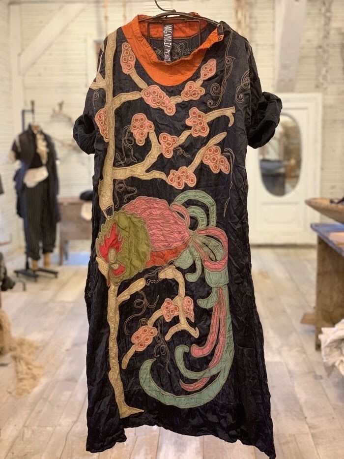 a closeup shot of a black dress hanging on a wooden hanger. the dress features intricate embroidery of a bird perched on a flowering branch, done in muted tones of pinks, greens, and golds against the dark fabric. the dress is made of a crinkled, possibly silk material, and has a relaxed fit with short sleeves and a high neckline. the background is a rustic setting of a clothing store, with wooden floors and other clothing items visible. the overall style evokes a bohemian, vintage, or artisan feel.