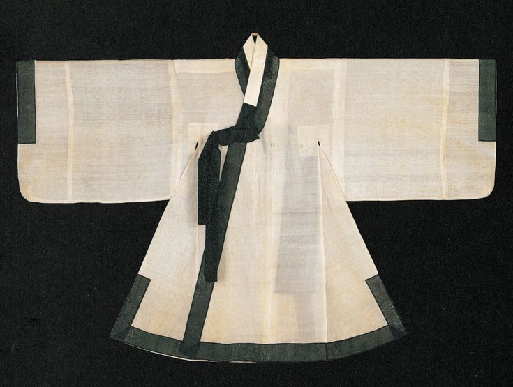 a full view of a hanbok garment laid out on a dark surface. the hanbok is mostly white with black trim on the sleeves and the triangular bottom portion. black ribbons are draped across the front. the sleeves are spread wide, and the overall design of the garment is visible.