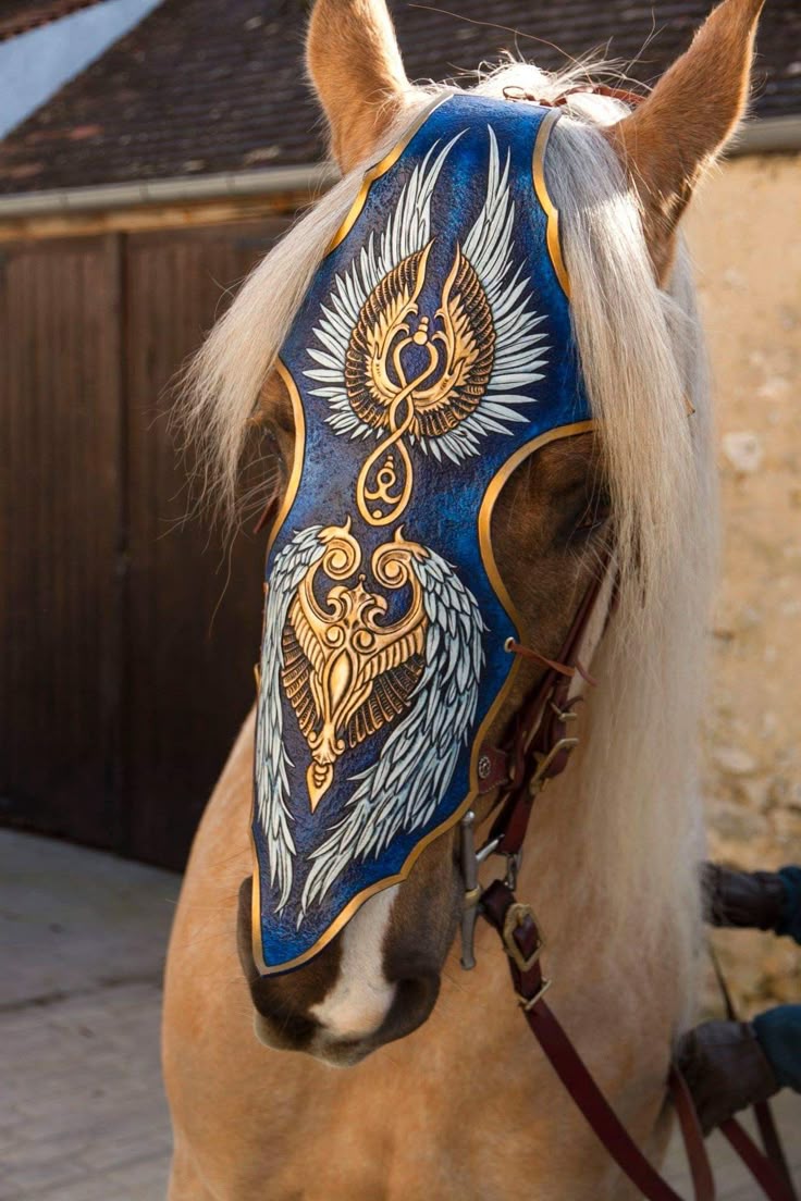 a closeup shows a lightbrown horse wearing an elaborate, blue face covering. the covering is adorned with gold and white designs, including images of winged creatures and intricate patterns. a bridle is visible under the covering, with a dark red rein. the horse has a white blaze on its nose and a lightcolored mane. the background features a building and some foliage.