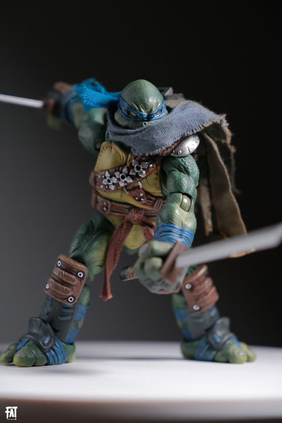 a studio shot of a teenage mutant ninja turtles leonardo action figure. the figure is highly stylized and detailed, presented in a postapocalyptic aesthetic. leonardo is holding two katana swords, his signature weapons. he is wearing his blue bandana and a rugged, worn outfit. the background is dark, which highlights the figure. the image is welllit and focused.