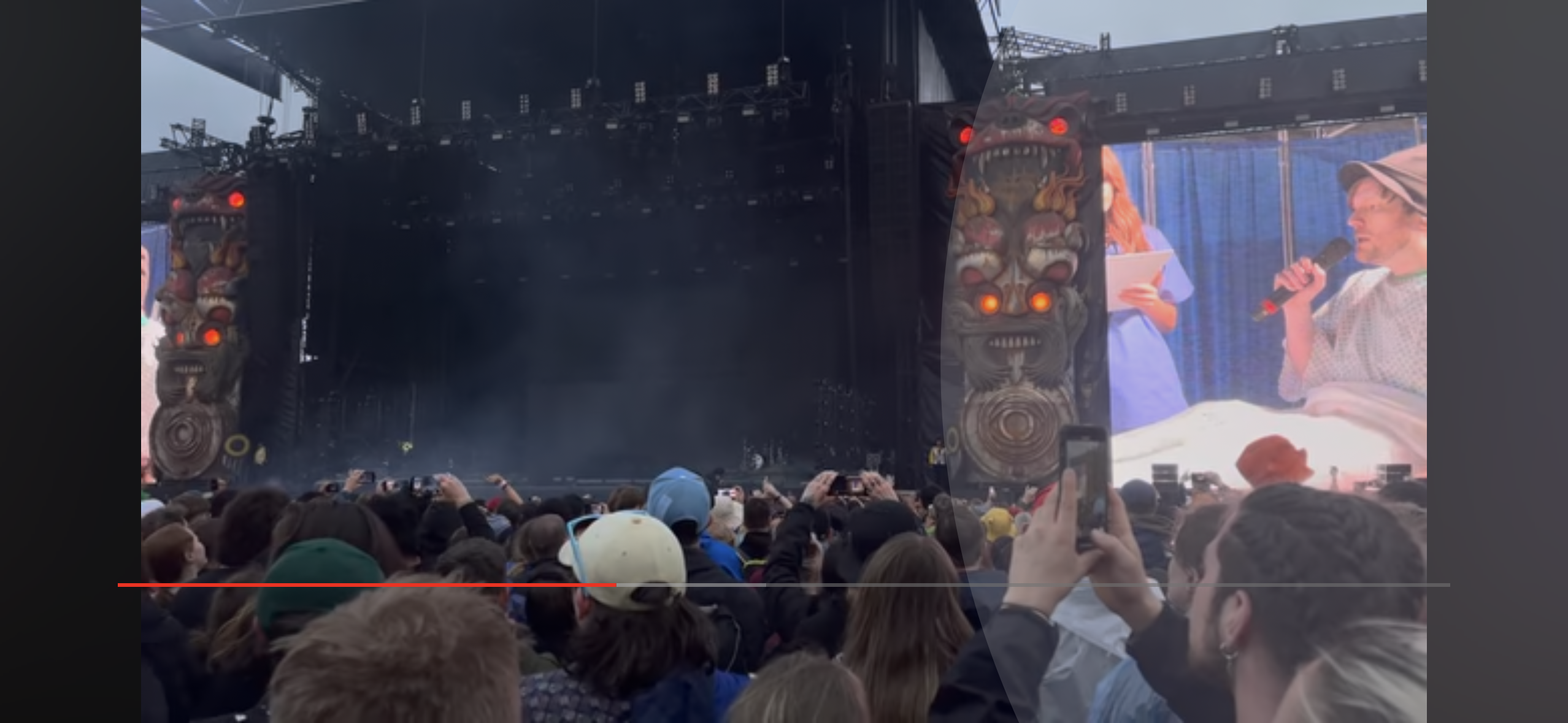 this is a photo of a concert from the perspective of a member of the audience. the audience is crowded together looking at the stage, which is lit with many lights, and behind the stage is a giant dark structure that appears to be a totem pole.  the image is captured during the night at what appears to be a summer music festival.