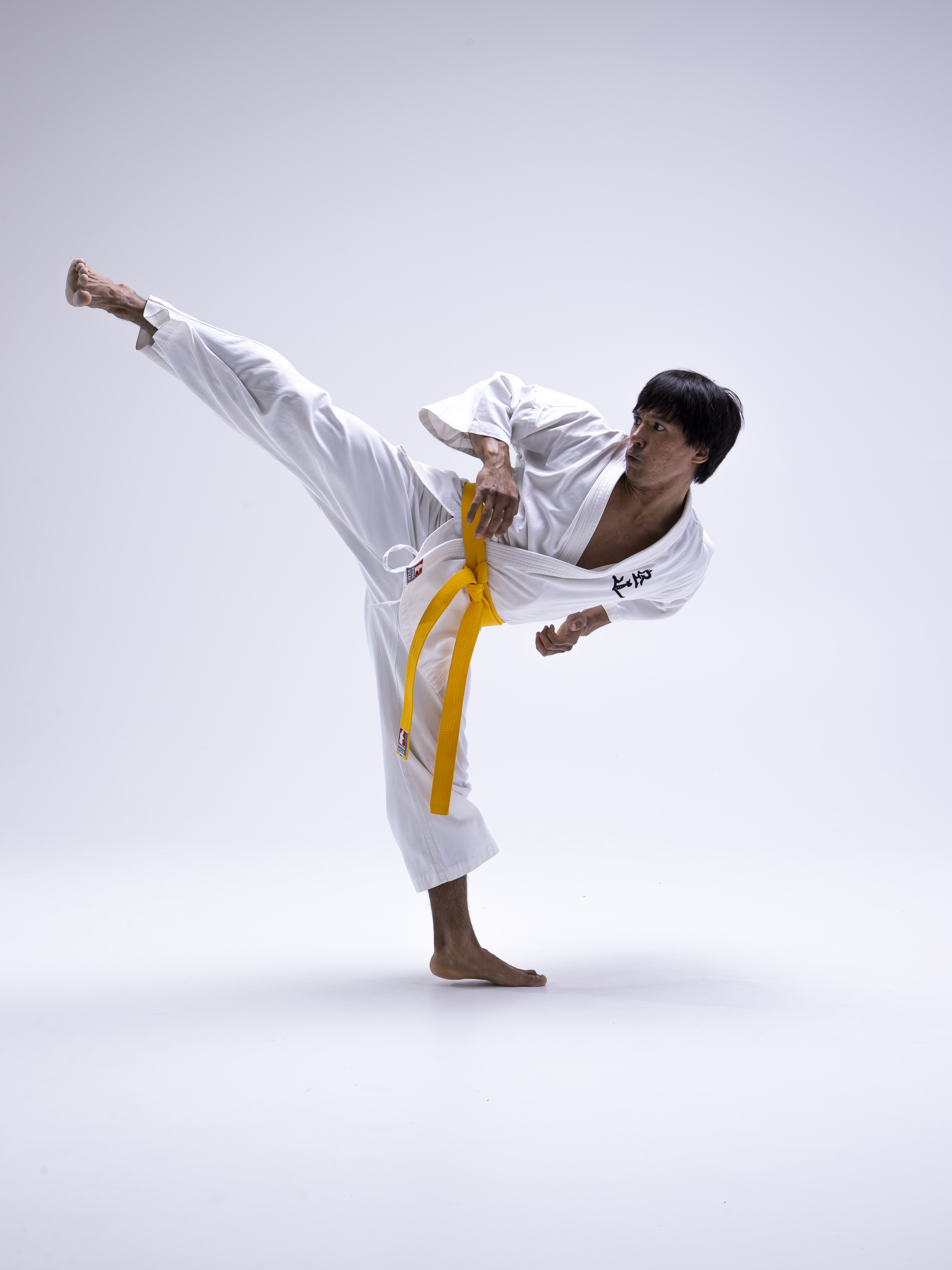 a man in a white karate gi with a yellow belt is performing a kick in a studio setting. he is barefoot and striking an action pose. the image is a full body shot against a white background.