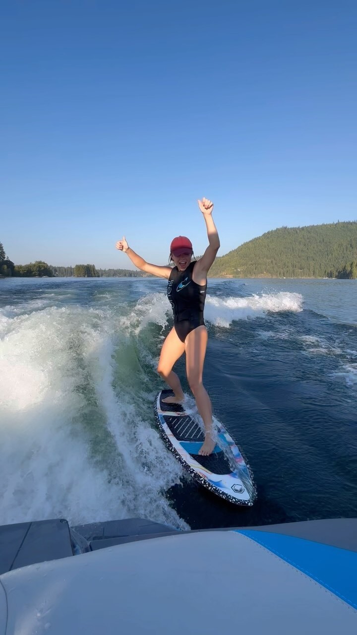 a woman wake surfing on a lake. she is wearing a black swimsuit and a red hat. she is smiling and has her arms raised in the air. the lake is surrounded by mountains and the sky is blue. the woman is riding a wave created by a boat. she is having fun and looks very happy.