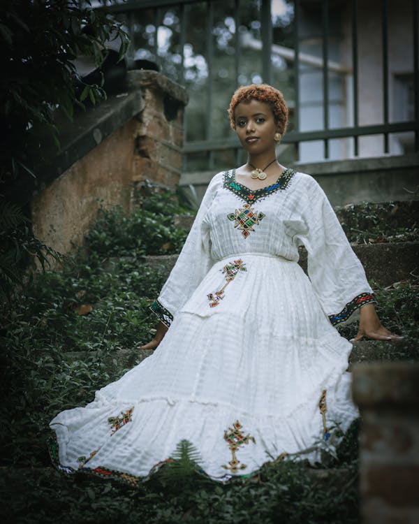 a young woman with short, reddishbrown hair is sitting on stone steps in an outdoor setting. she is wearing a white, longsleeved dress with colorful embroidery on the neckline, sleeves, and skirt. she also has a necklace and earrings. the steps are overgrown with greenery, and a stone structure and a metal gate are visible in the background. the lighting is soft and diffused, creating a serene and slightly melancholic atmosphere.