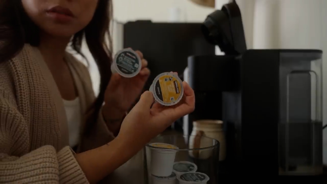 a woman is holding two coffee pods in her hands, deciding which one to use. she is standing in a kitchen with a coffee maker in the background. there is a container of coffee pods on the counter next to the coffee maker.