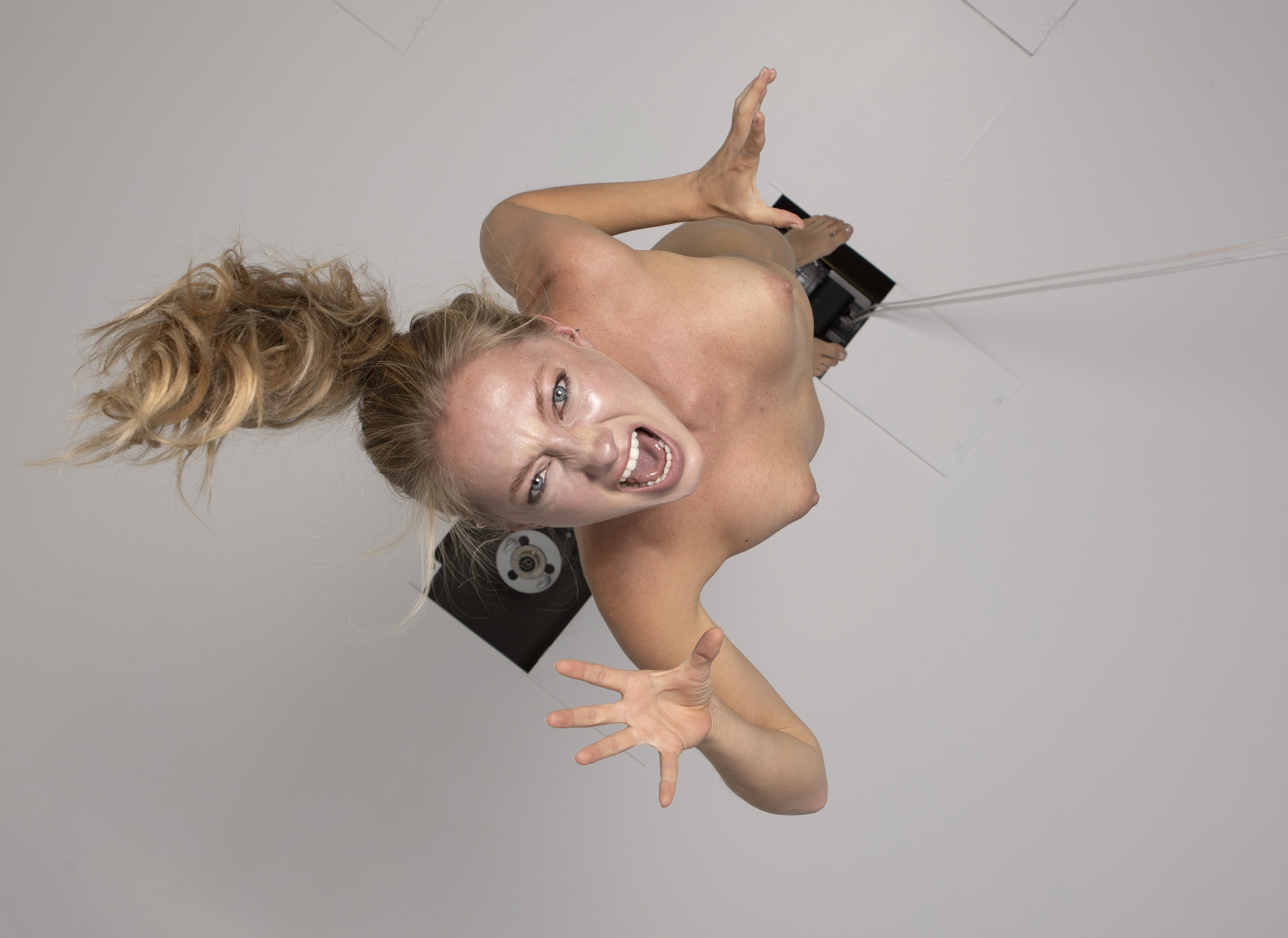 an overhead shot captures a topless woman with blonde hair pulled back in a ponytail, screaming with an angry expression. her hand is outstretched towards the camera. she is standing on some kind of studio setup. the perspective is from directly above, looking down at her.