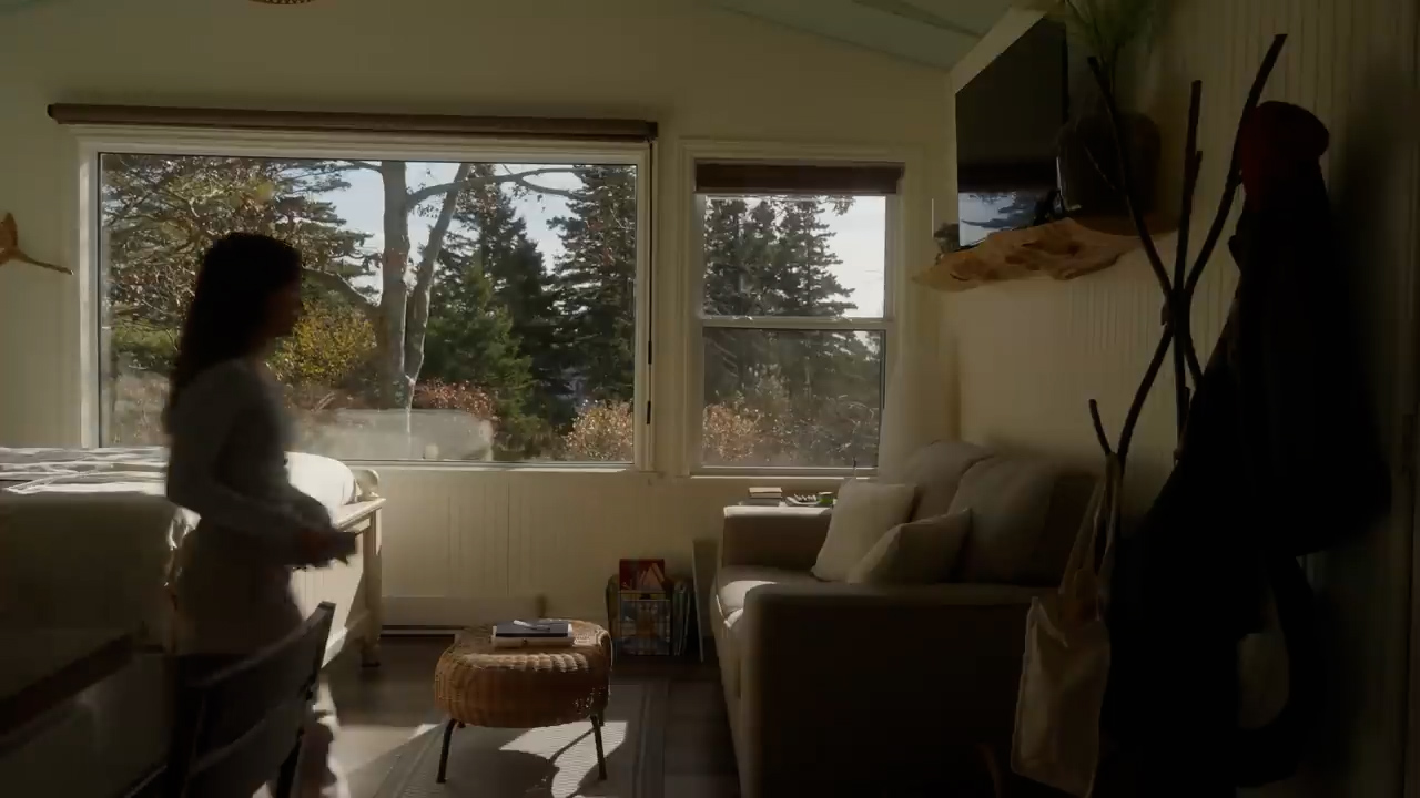 a woman is walking in a living room. there is a couch, a window, a bed, and a coffee table in the room. the room is lit by natural light.